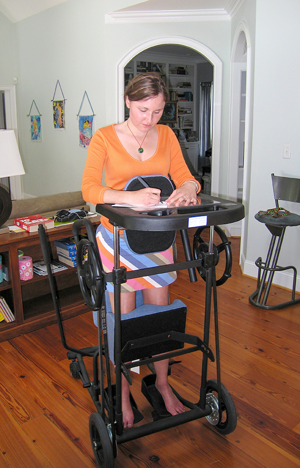 Woman standing in an assistive wheelchair and writing on an attached desktop.