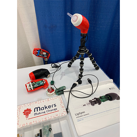 A mouth operated joystick sits on a tabletop tripod. Next to it is a parts kit from Makers Making Change, and an assembly manual and a second, partially assembled 3-D printed LipSync.
