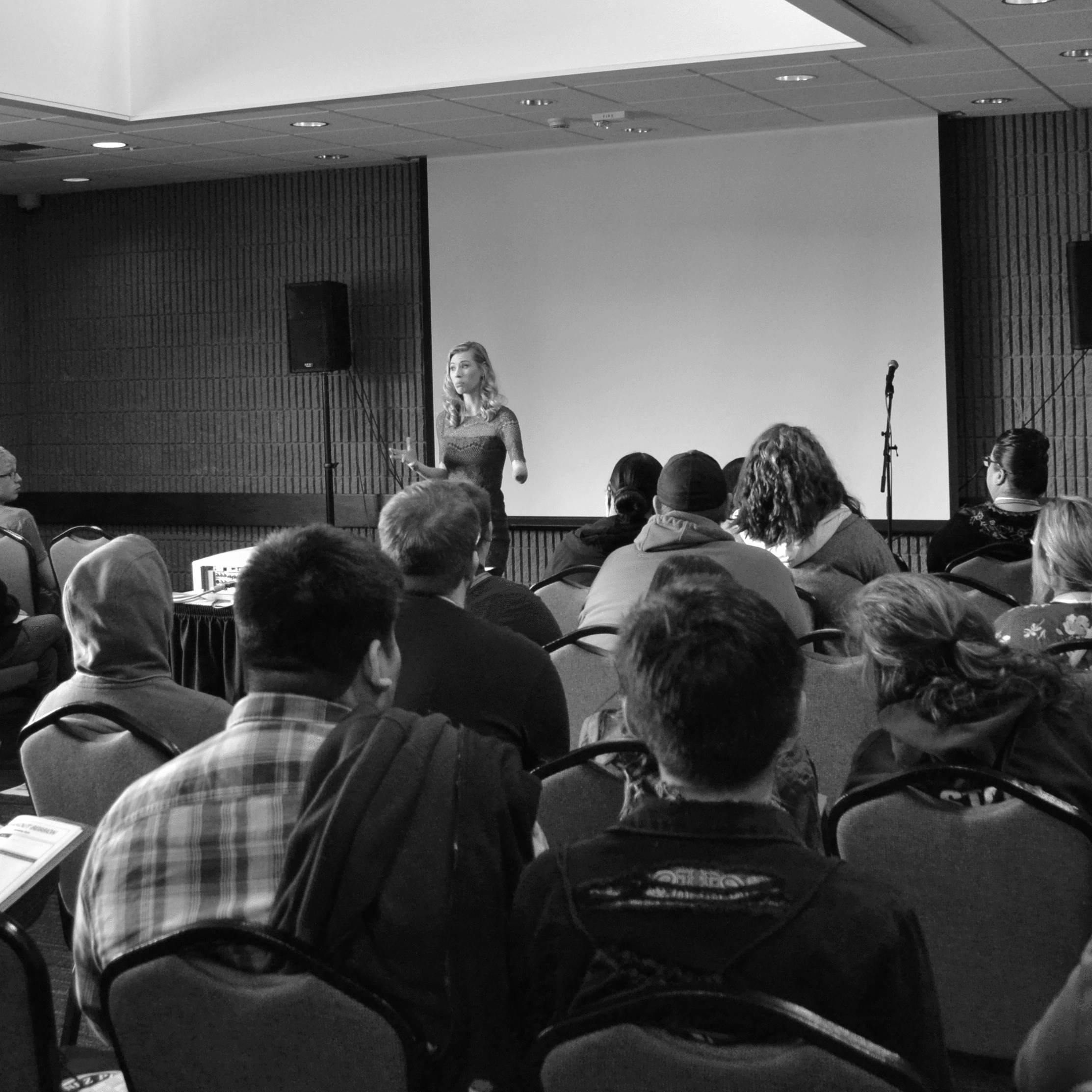 A woman with one arm standing in front of a crowd in a conference room. She is giving a presentation. She is wearing a red dress and has mid-length blonde hair. Everyone in the room is watching her thoughtfully.
