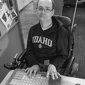 A man with a disability is using an adaptive keyboard and wheelchair as he sits at his workdesk. He is looking at the camera with a slight smile. He is wearing glasses and a University of Idaho sweatshirt.